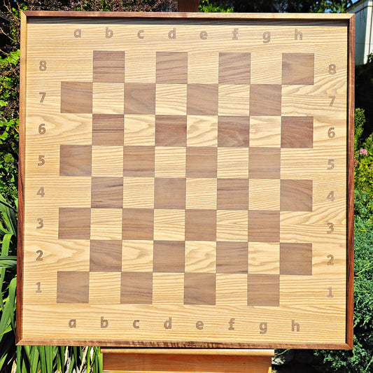 Front view of the empty ChessboArt 800 wooden chessboard bathed in natural sunlight. The image showcases the striking wood grain of the light heartwood ash and dark American walnut squares, which flow seamlessly into the coordinate margins without any inner separating border.