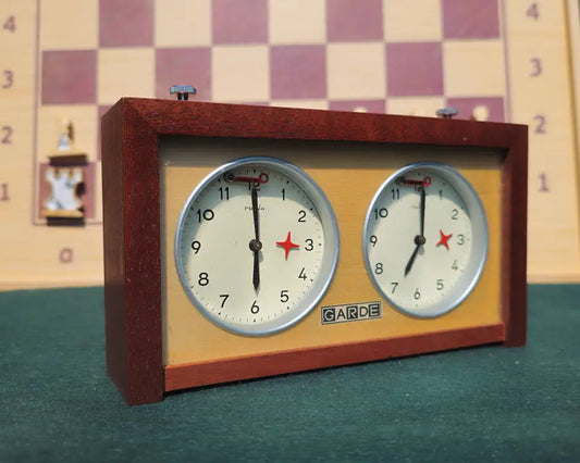 A pristine vintage UMF Ruhla Gardé 2nd generation mechanical chess clock from the mid-1960s (Made in GDR), viewed from the front-right on a green felt surface. The clock features a solid dark wood case and crisp white dials with the distinct 'GARDE' logo. A blurred wooden chessboard and pieces, including a Rook and Pawn, are in the background.