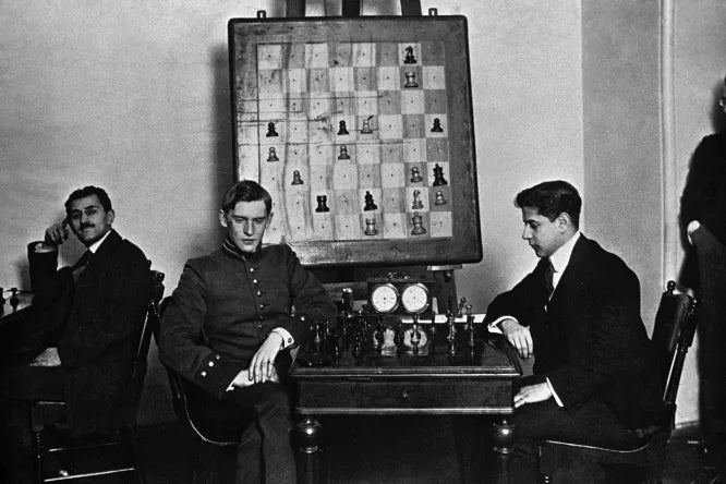Alekhine and Capablanca playing a game at the table, in the background a large demonstration chessboard on the wall (ca. 1913).