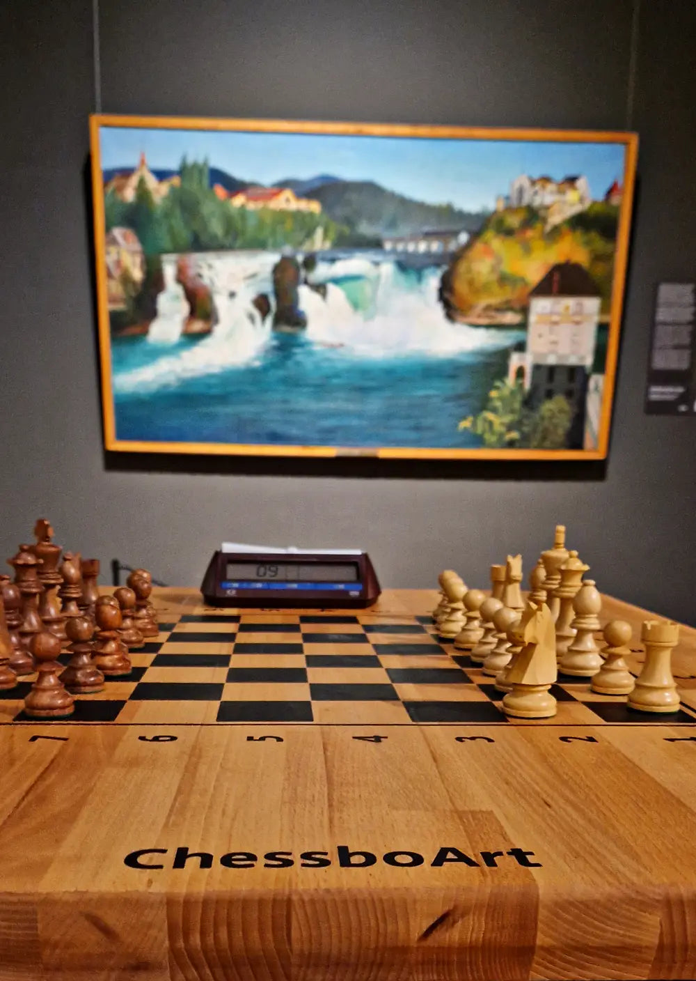 Luxury wooden chessboard by ChessboArt displayed in an art gallery, with a chess clock and a painting in the background.