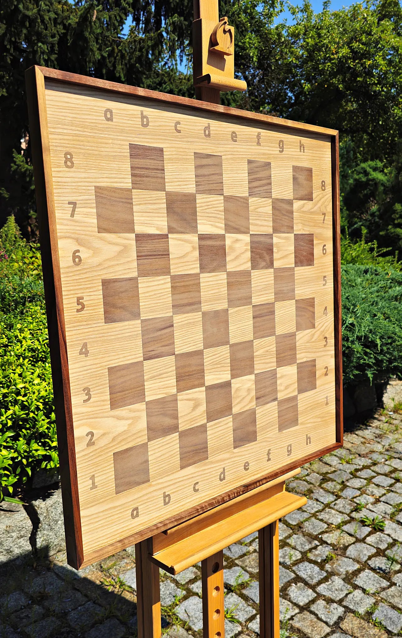 Angled perspective of the empty ChessboArt 800 large wooden chessboard displayed on an outdoor easel. This side view highlights the thick, solid American walnut frame and the unique seamless transition from the playing squares to the coordinate margins.