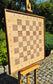 Angled perspective of the empty ChessboArt 800 large wooden chessboard displayed on an outdoor easel. This side view highlights the thick, solid American walnut frame and the unique seamless transition from the playing squares to the coordinate margins.