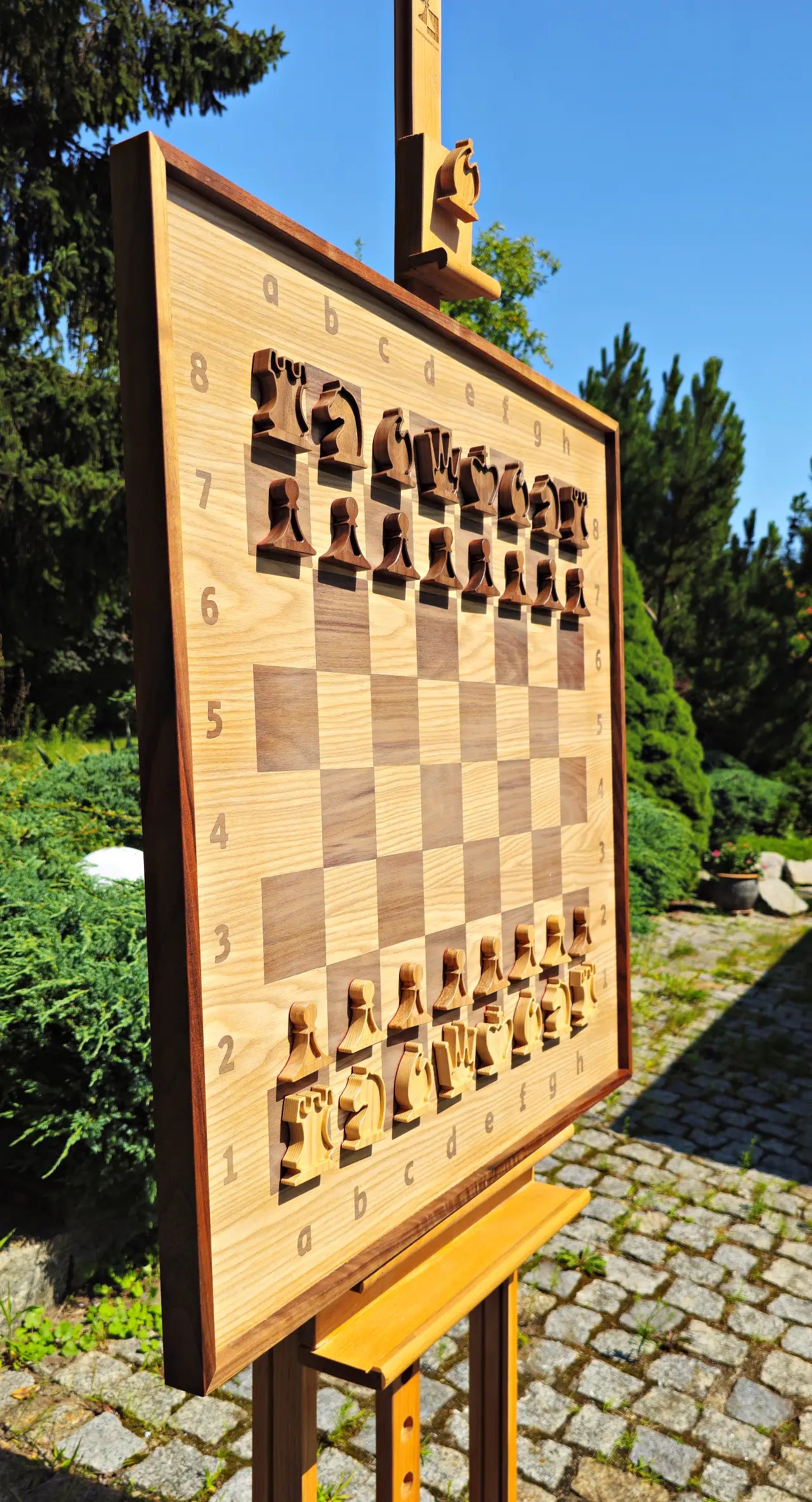 Side perspective of the large ChessboArt 800 wall chessboard displayed on an outdoor easel. The 800x800mm borderless board, crafted from heartwood ash and American walnut, is set up with custom flat solid wood chess pieces. The angled view highlights the board's thick walnut frame and modern seamless design.
