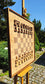 Side perspective of the large ChessboArt 800 wall chessboard displayed on an outdoor easel. The 800x800mm borderless board, crafted from heartwood ash and American walnut, is set up with custom flat solid wood chess pieces. The angled view highlights the board's thick walnut frame and modern seamless design.