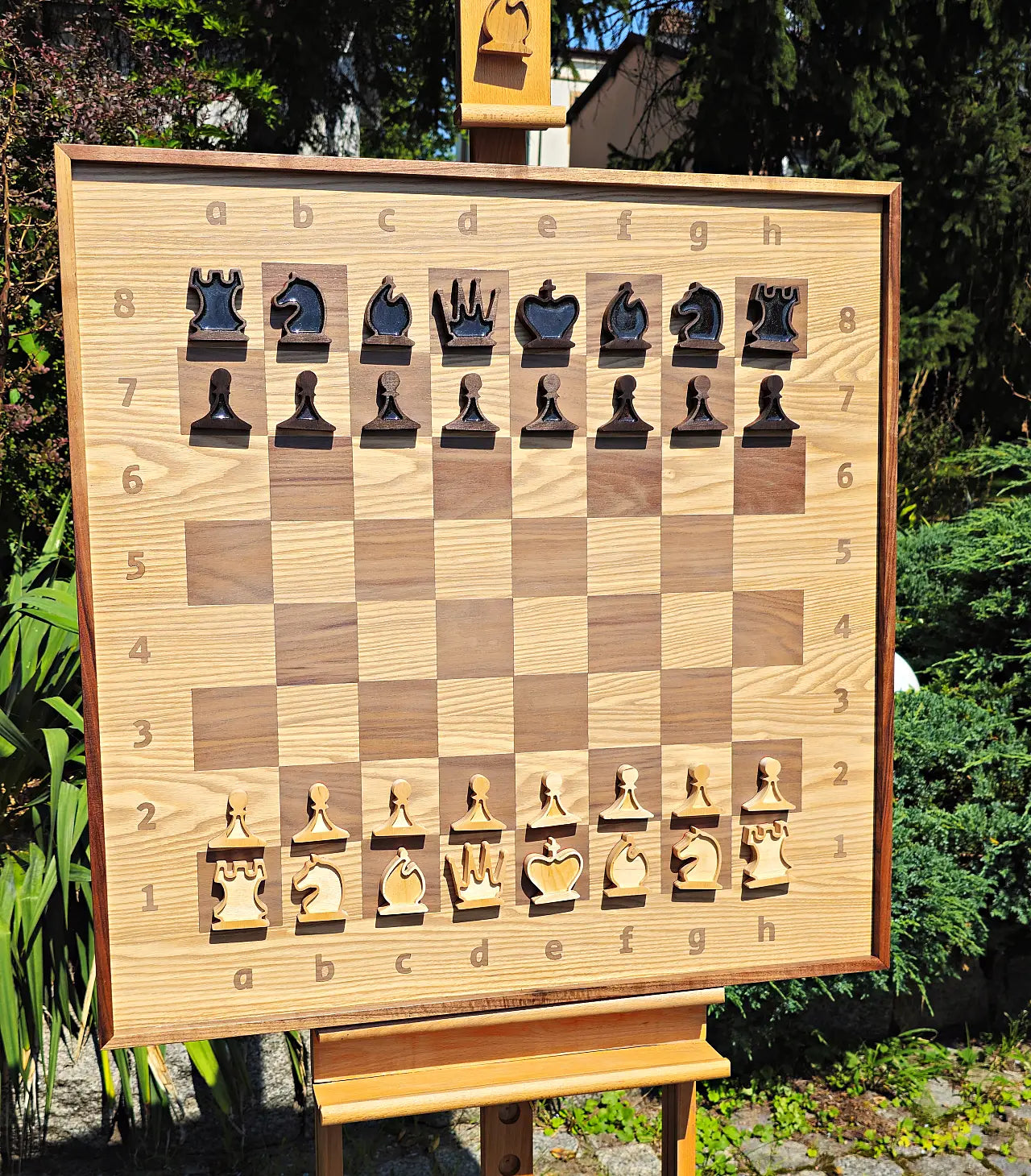 The ChessboArt 800 large wall chessboard displayed on a wooden easel. This setup features unique plywood wall chess pieces. The black pieces are beautifully filled with glossy black resin, while the white pieces remain open and unfilled. The 800x800mm board showcases a borderless design made of ash and American walnut.