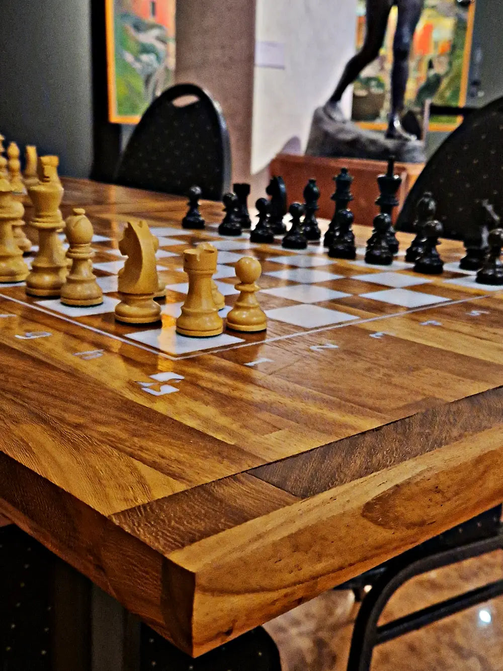 A full view of a handcrafted ChessboArt bughouse chess table made from Iroko wood, with both white and black chess pieces set up for a game. The warm wooden surface and a subtle reflection of the background highlight the table's premium craftsmanship.