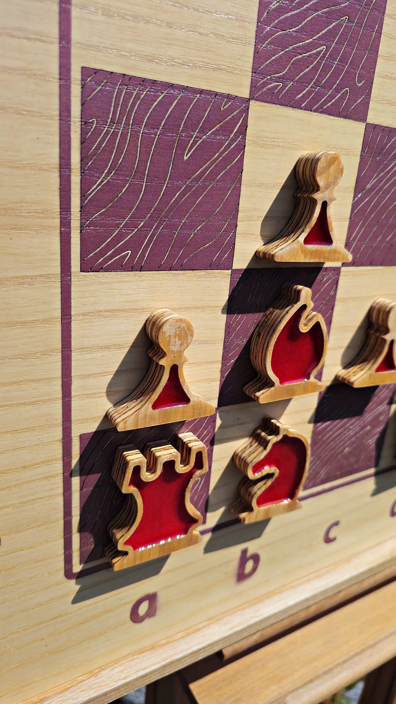 Angled close-up of the ChessboINK wall-mounted chessboard made of pink-painted ash wood, featuring unique flat wooden chess pieces with vibrant red epoxy resin fillings casting shadows in the sunlight.