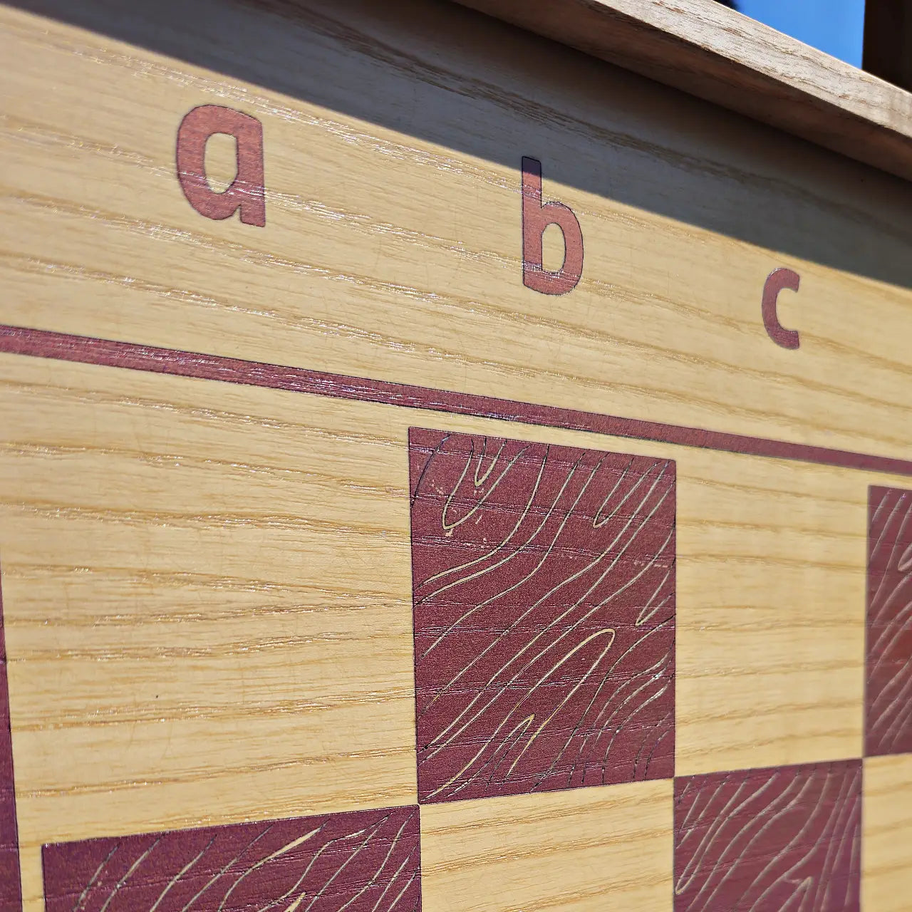 Close-up detail of the ChessboINK ash wood chessboard, showcasing the textured natural wood grain beneath the pink painted squares and the algebraic notation letters a, b, and c.
