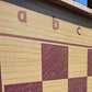 Close-up detail of the ChessboINK ash wood chessboard, showcasing the textured natural wood grain beneath the pink painted squares and the algebraic notation letters a, b, and c.