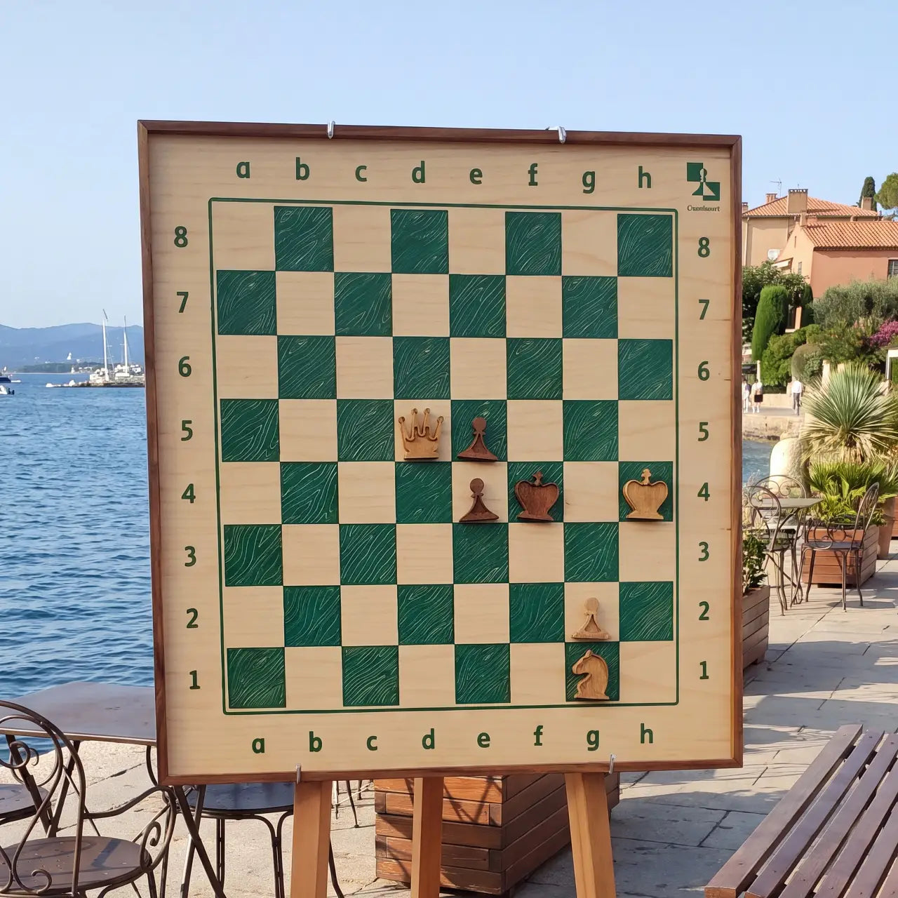 ChessboINK wall chess board in ash with hand-painted green fields and a walnut frame, shown with a few pieces on a stand by the sea