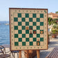 ChessboINK wall chess board in ash with hand-painted green fields and a walnut frame, shown with a few pieces on a stand by the sea