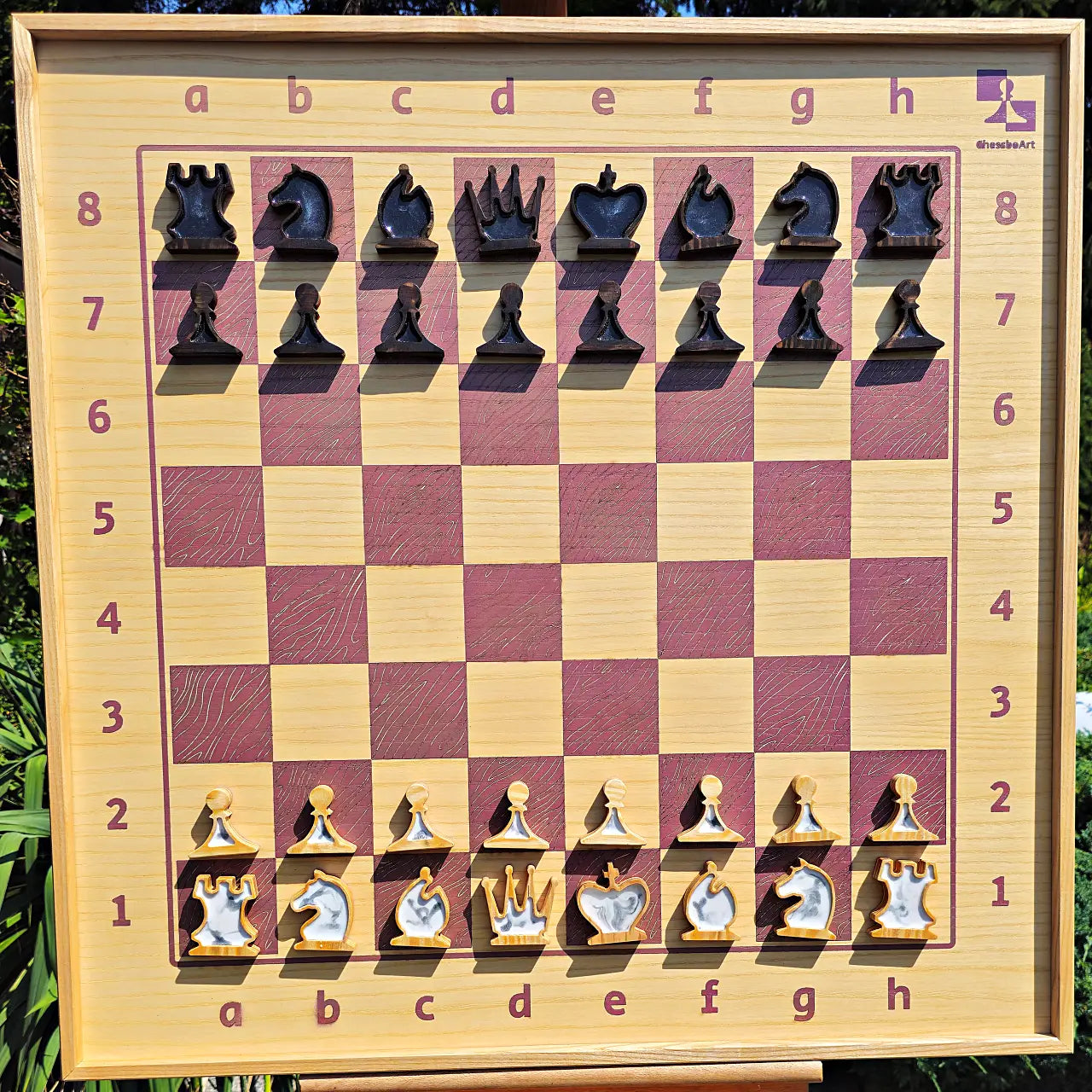 Front view of the complete ChessboINK wall-mounted chessboard made of pink and natural ash wood, featuring a full set of flat wooden chess pieces with black and white pearl epoxy resin fillings set up in their starting positions.