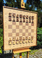 The ChessboArt 800 large wooden chess board displayed on an easel outdoors. Crafted from American walnut and heartwood ash, this 800x800mm board features a unique minimalist design with a seamless transition and no inner border separating the squares from the coordinate margins. Set up with solid wood flat profile pieces.