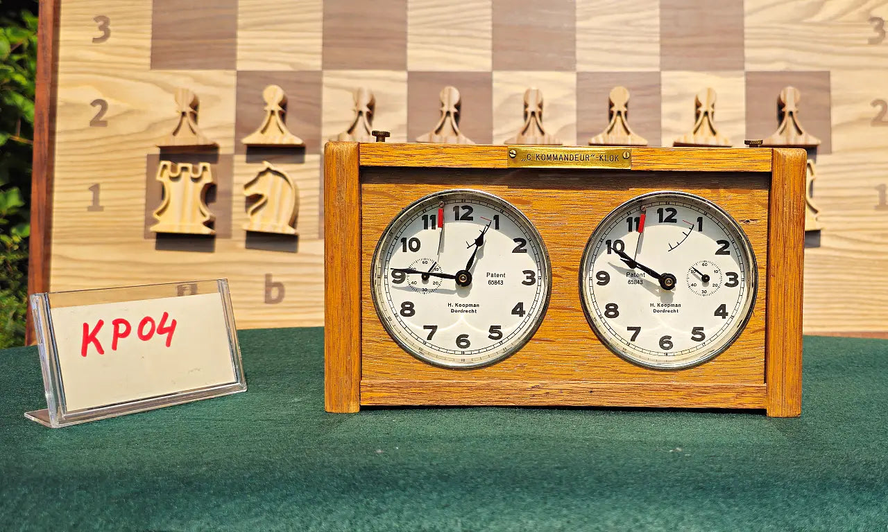 Front view of a classic mid-century wooden chess timer. This collectible Koopman clock from the 1960s, model KP04, features twin analog dials and a brass nameplate, sitting on a green game table.