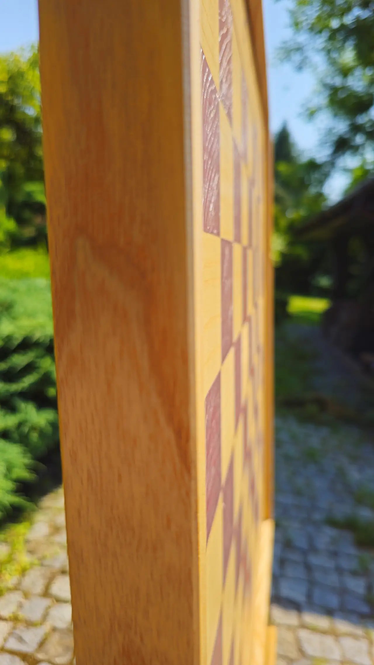 Side profile view of the ChessboINK wall-mounted chessboard, focusing on the solid ash wood frame thickness and smooth natural wood finish against a blurred outdoor background.