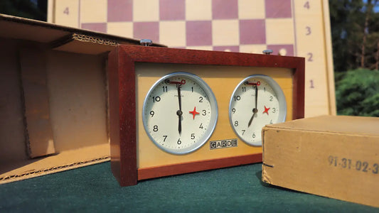 Mint condition vintage UMF Ruhla Gardé 2nd generation mechanical chess clock from the 1960s, displayed alongside its rare original cardboard factory box with a visible reference stamp. The dark wood timer sits on green felt with a softly blurred chessboard behind it.