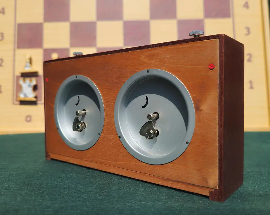 Rear view of the mint condition vintage UMF Ruhla Gardé 2nd generation mechanical chess clock. The solid dark wood case shows the dual integrated wind-up keys and setting knobs on silver back-plates. The clock rests on green felt with a softly blurred wooden chessboard in the background, highlighting the authentic dual mechanism.