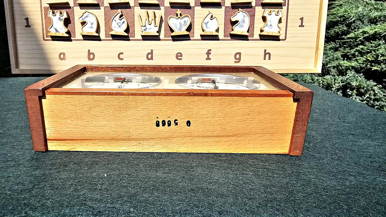 Bottom view of the vintage UMF Ruhla Gardé 2nd generation mechanical chess clock, revealing the original black factory numbers stamped on the light wood base. The clock is resting flat on a green felt surface, with a wooden chessboard and epoxy pieces subtly visible in the background.