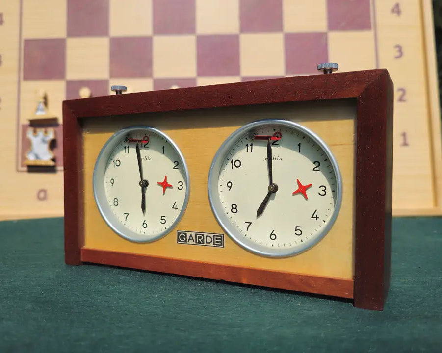 Front-left angle view of a mint condition vintage UMF Ruhla Gardé 2nd generation chess clock from the 1960s. Resting on a green felt surface, the mechanical GDR timer displays its solid dark wood case, clear dual dials with red flags, and a softly blurred wooden chessboard in the background.