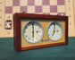 A pristine vintage UMF Ruhla Gardé 2nd generation mechanical chess clock from the mid-1960s (Made in GDR), viewed from the front-right on a green felt surface. The clock features a solid dark wood case and crisp white dials with the distinct 'GARDE' logo. A blurred wooden chessboard and pieces, including a Rook and Pawn, are in the background.